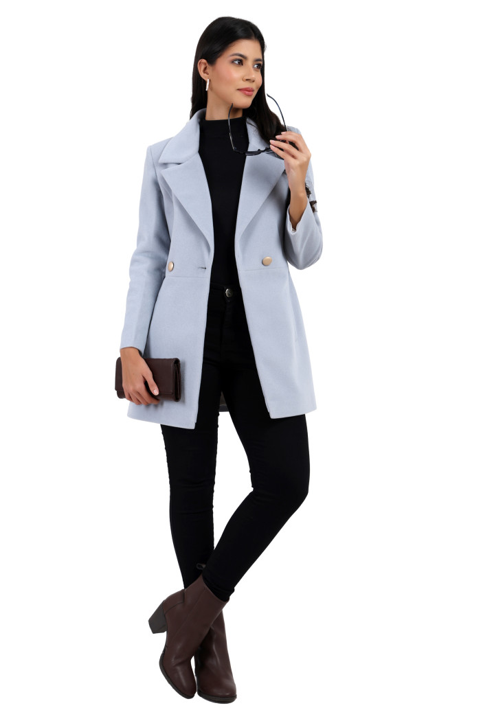 A woman in a standing pose is wearing Trufit’s double-breasted velvet coat in sky blue with a lapel collar, side pockets, and a button closure in black jeans while holding shades from her left hand and a clutch from her right hand.